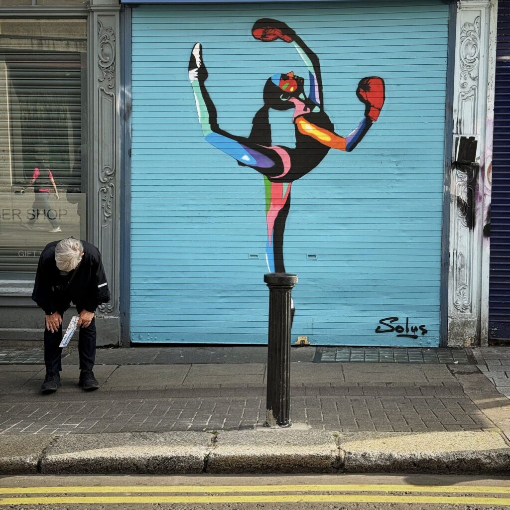 Dublin street photograph of a colourful Solus mural on a shutter with a seated
man, Brendan Ó Sé workshop